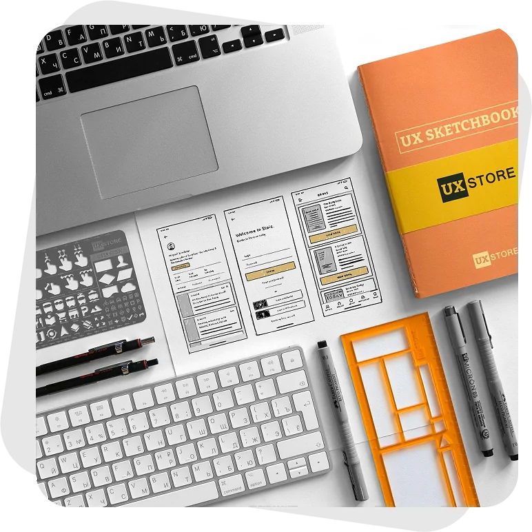 A designer's desk from above: a laptop, a keyboard, hand-drawn mobile wireframes, a UX sketchbook box, drawing stencils, technical pens and an orange layout ruler